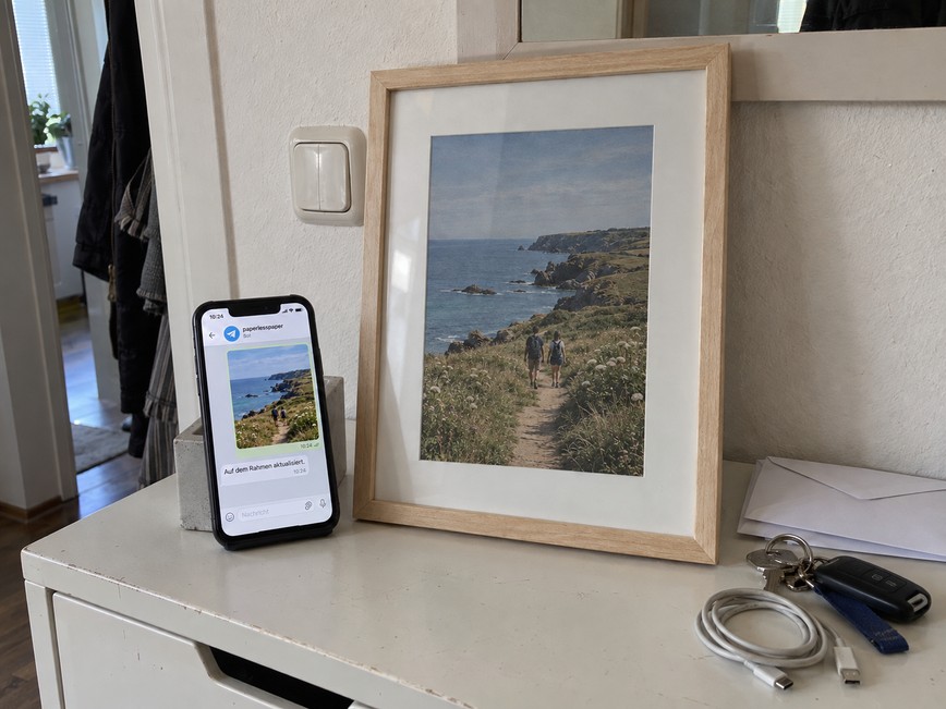 OpenPaper 13 und Handy auf einer Kommode zeigen dasselbe per Telegram gesendete Küstenfoto.